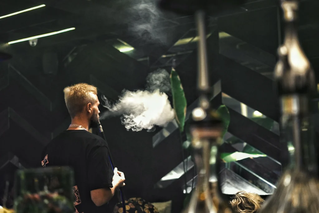 A man smoothly exhaling a thick cloud of smoke in a stylish, dimly lit lounge, representing the rewarding feeling of finally mastering a hookah for beginner session.