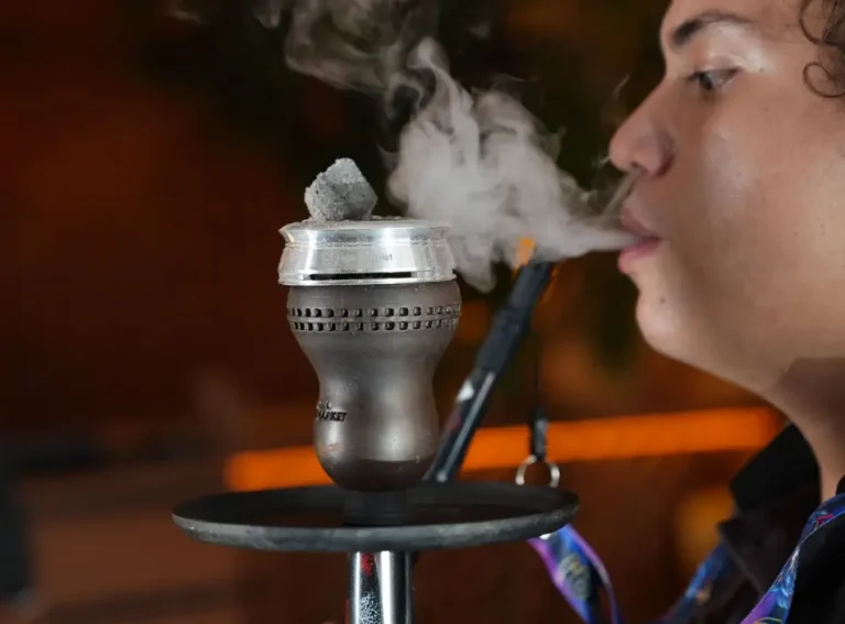 A person taking a smooth draw from a hookah equipped with a heat management device and a lit charcoal cube on top, showing how to control heat properly in a hookah for beginner.