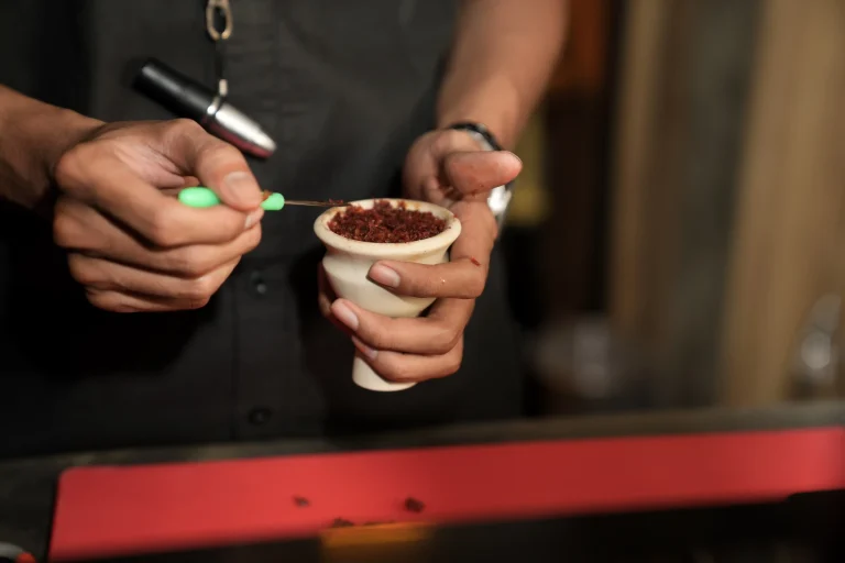 A close-up view of someone carefully packing red shisha tobacco into a white bowl using a small tool, illustrating the simple fluff pack method for a hookah for beginner.