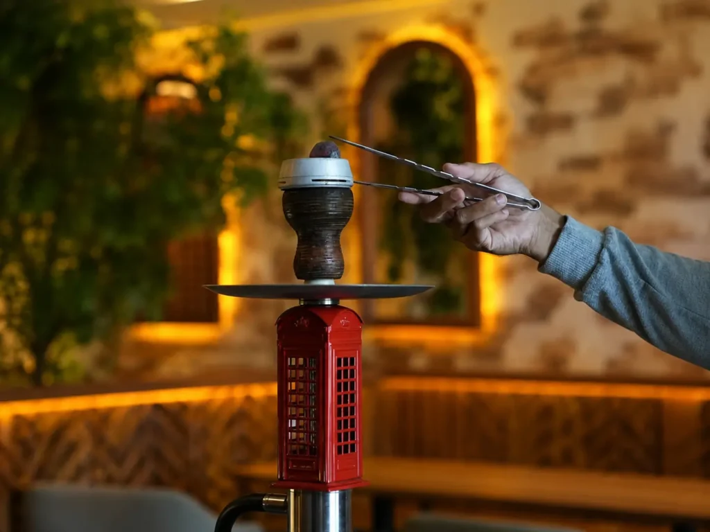 A close-up of a person using metal tongs to carefully place a piece of charcoal into a silver heat management device on top of a red phone booth-themed hookah, illustrating a key heat management step for any hookah for beginner setup.